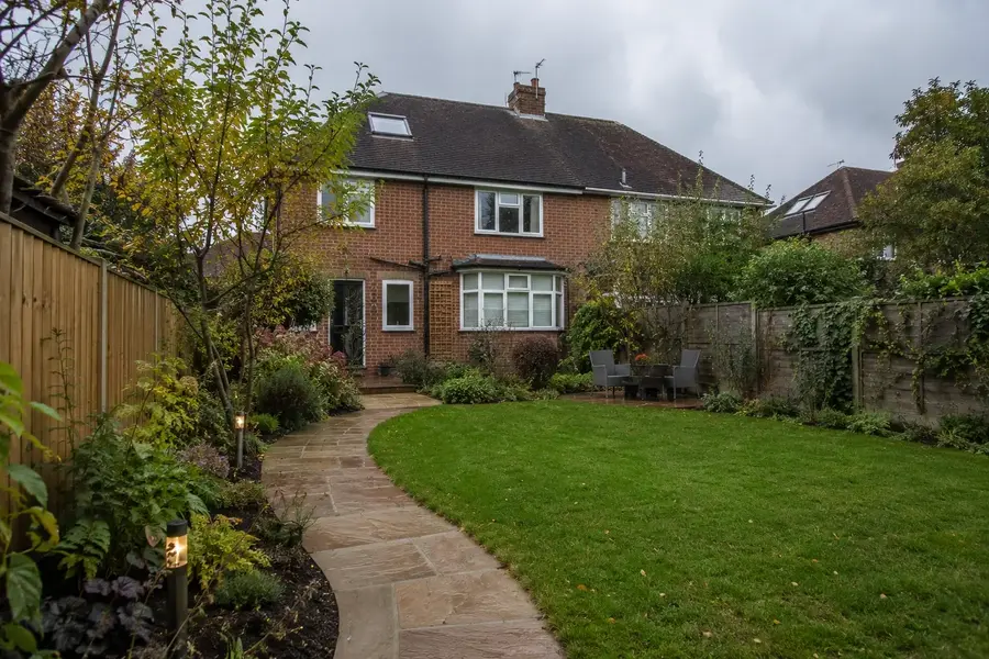 A curved sandstone path through a well-kept lawn, with layered planting and garden lighting outside a Surrey brick home