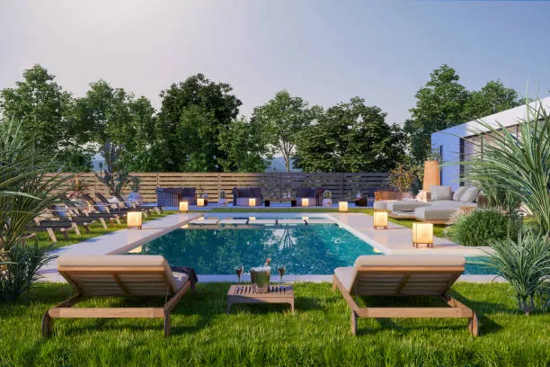 A modern landscaped garden featuring a rectangular swimming pool surrounded by a paved patio, with wooden lounge chairs on a lush lawn and decorative lantern lighting along the pool edge.