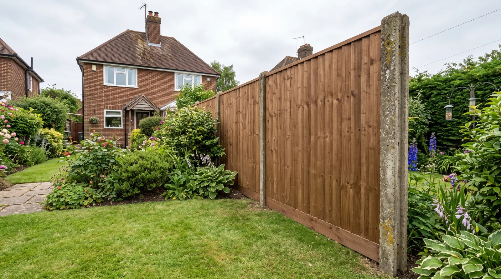 A new timber fence panel installed between older concrete slotted posts in a suburban garden, featuring a green lawn, mature plants, and a traditional red-brick house in the background.   
