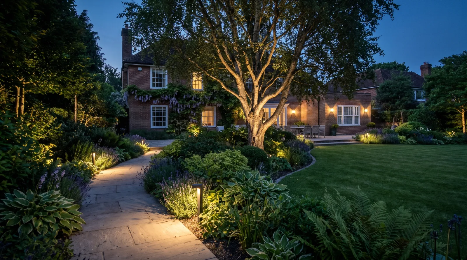Subtle garden lighting illuminating a path, specimen tree and planting borders in a landscaped Surrey garden at dusk.